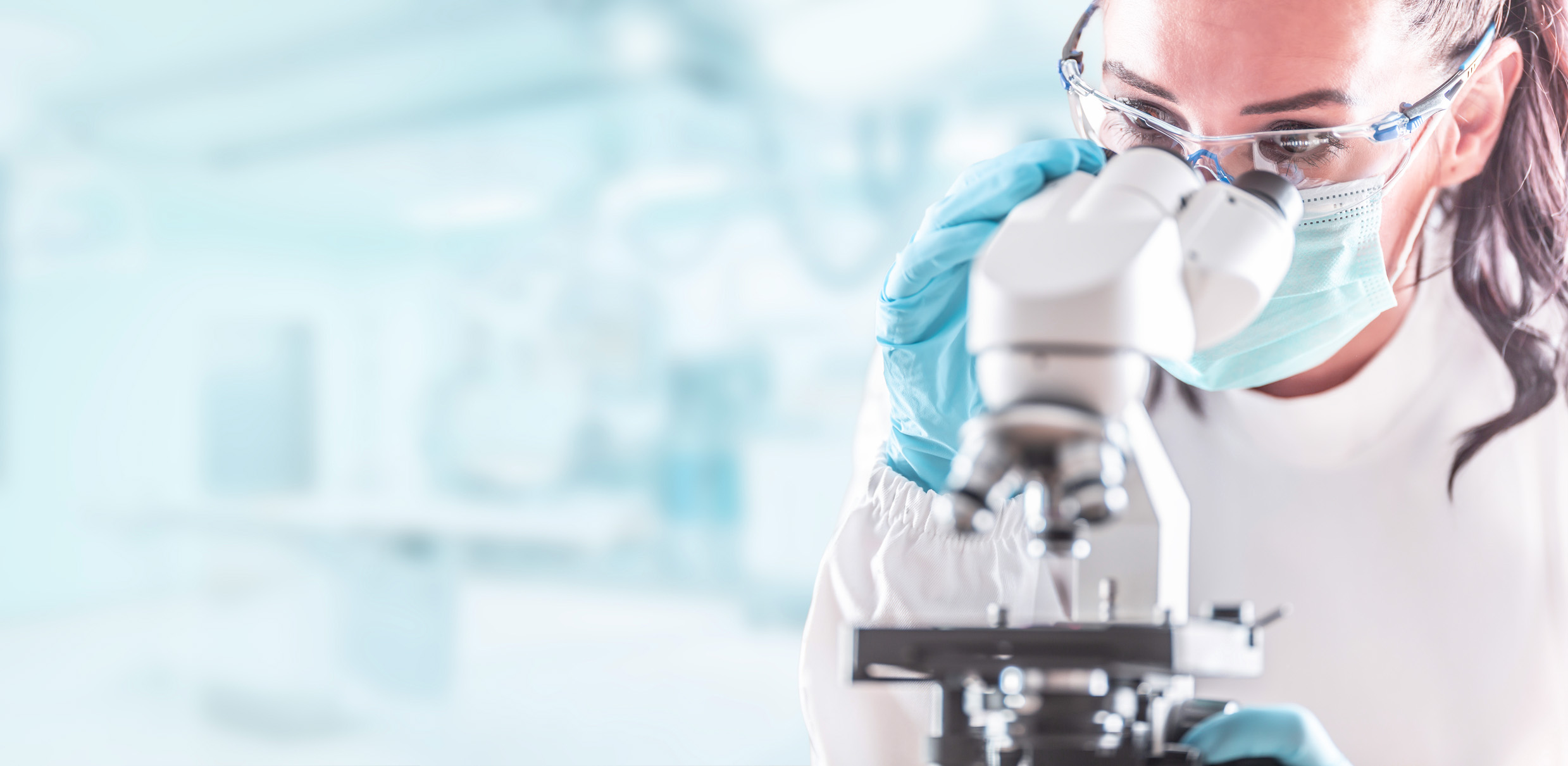 Female lab technician in protective glasses, gloves and face mask sits next to a microscope in laboratory.